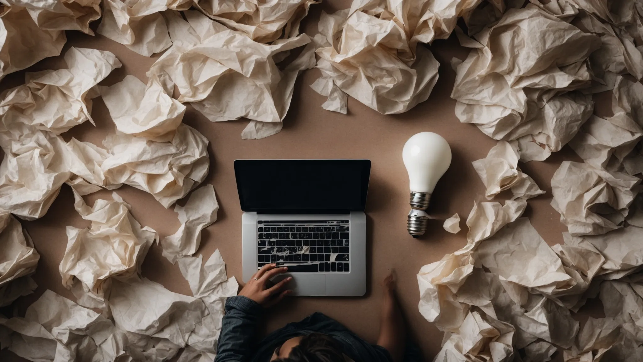 a person hovers over a laptop surrounded by crumpled paper and a light bulb—symbolizing the struggle between creative thinking and algorithmic strategies.