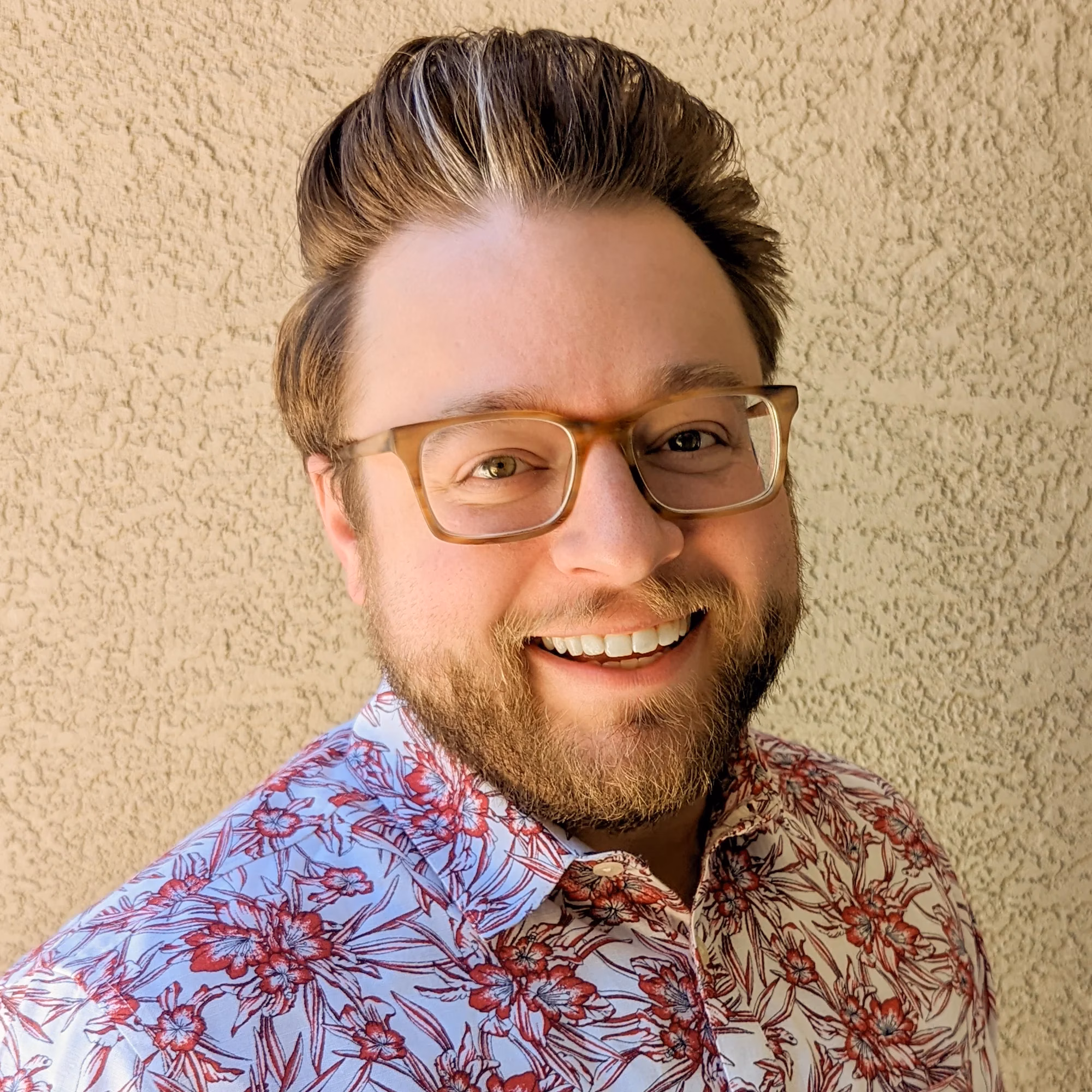 Smiling person with glasses wearing a floral shirt against a textured wall background.